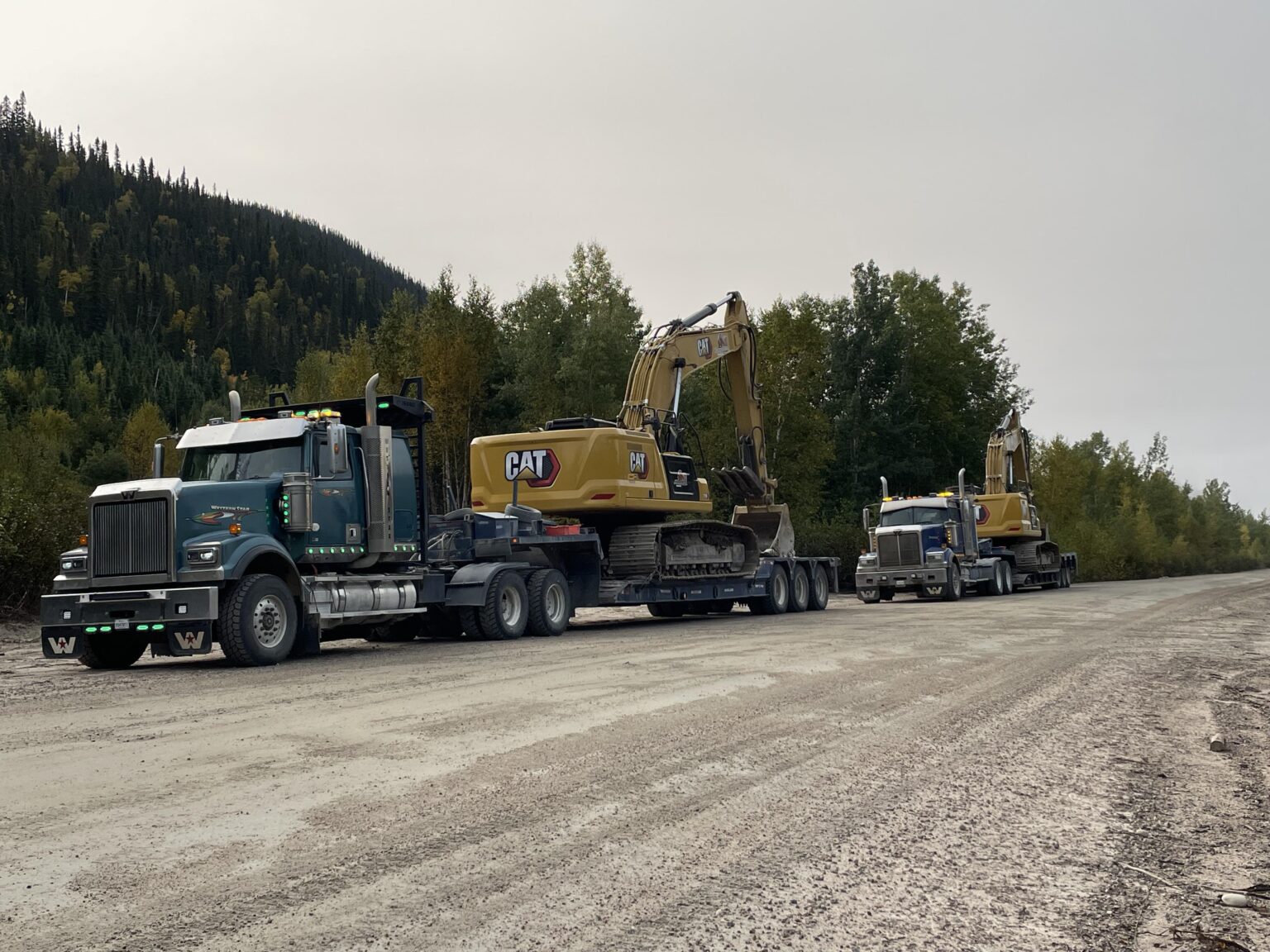Deux camions transportant une pelle mécanique sur un chemin de terre.